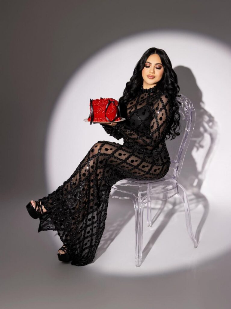 A woman in a black sequined sheer dress sits on a clear chair holding a red cake.
