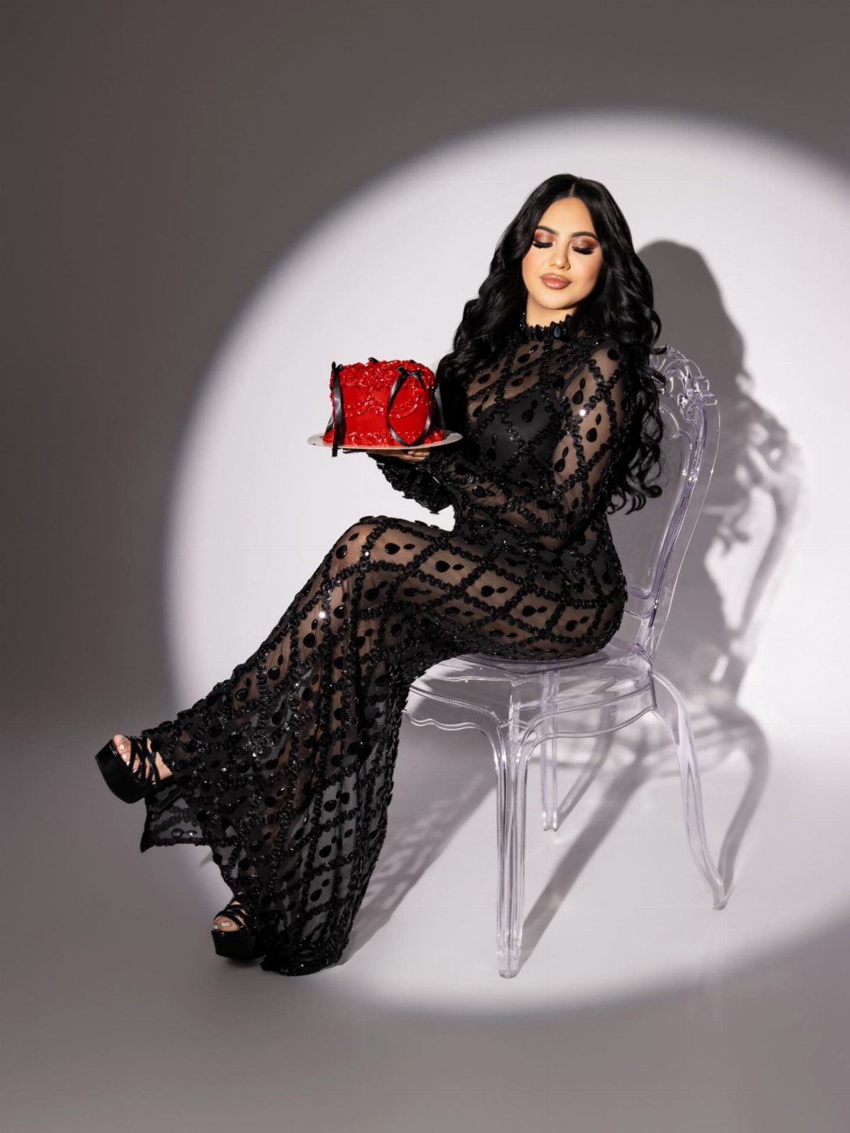 A woman in a black sequined sheer dress sits on a clear chair holding a red cake.