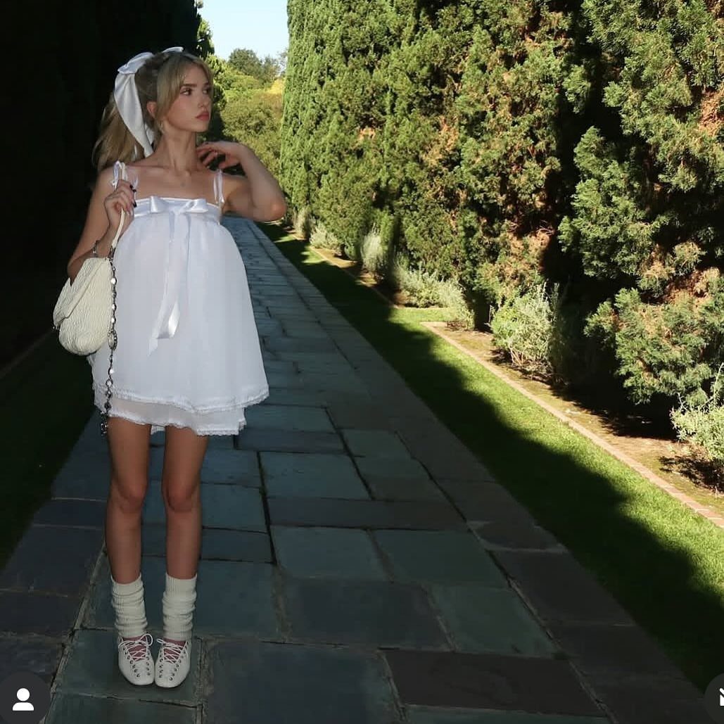 A woman in a white babydoll dress and leg warmers standing on a stone path.