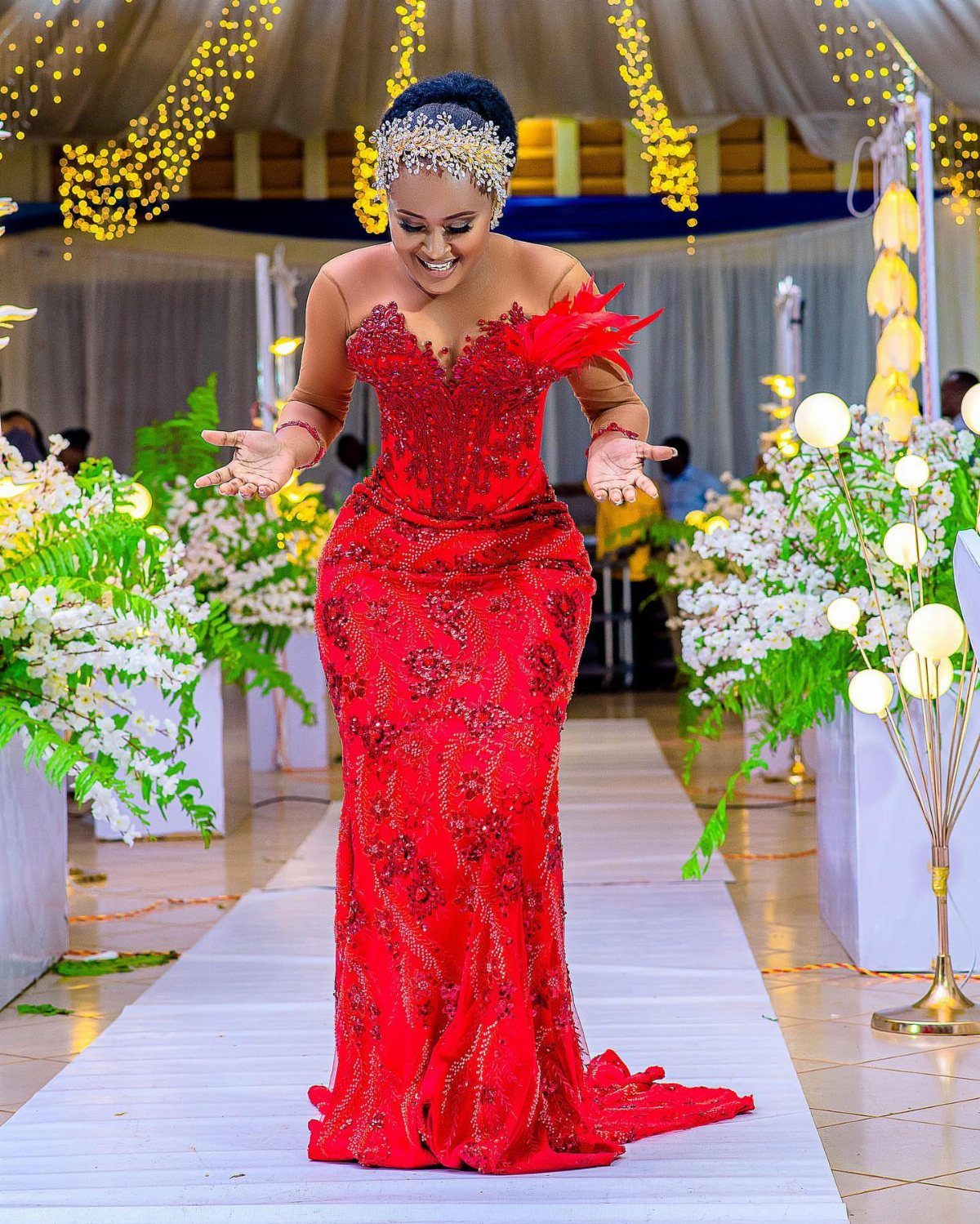 Woman in a red beaded mermaid dress walking down a decorated runway with white flowers.