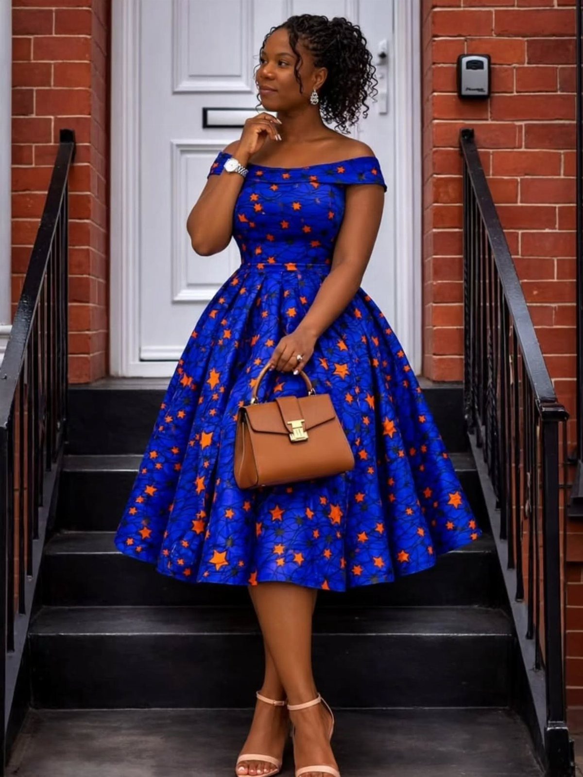 A woman posing in a blue and orange patterned structured midi dress holding a brown handbag.