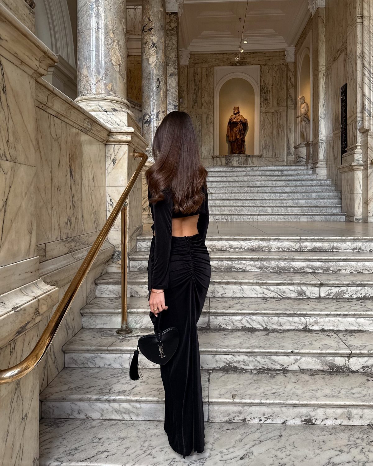 Woman in a long black backless velvet dress standing on a grand marble staircase.