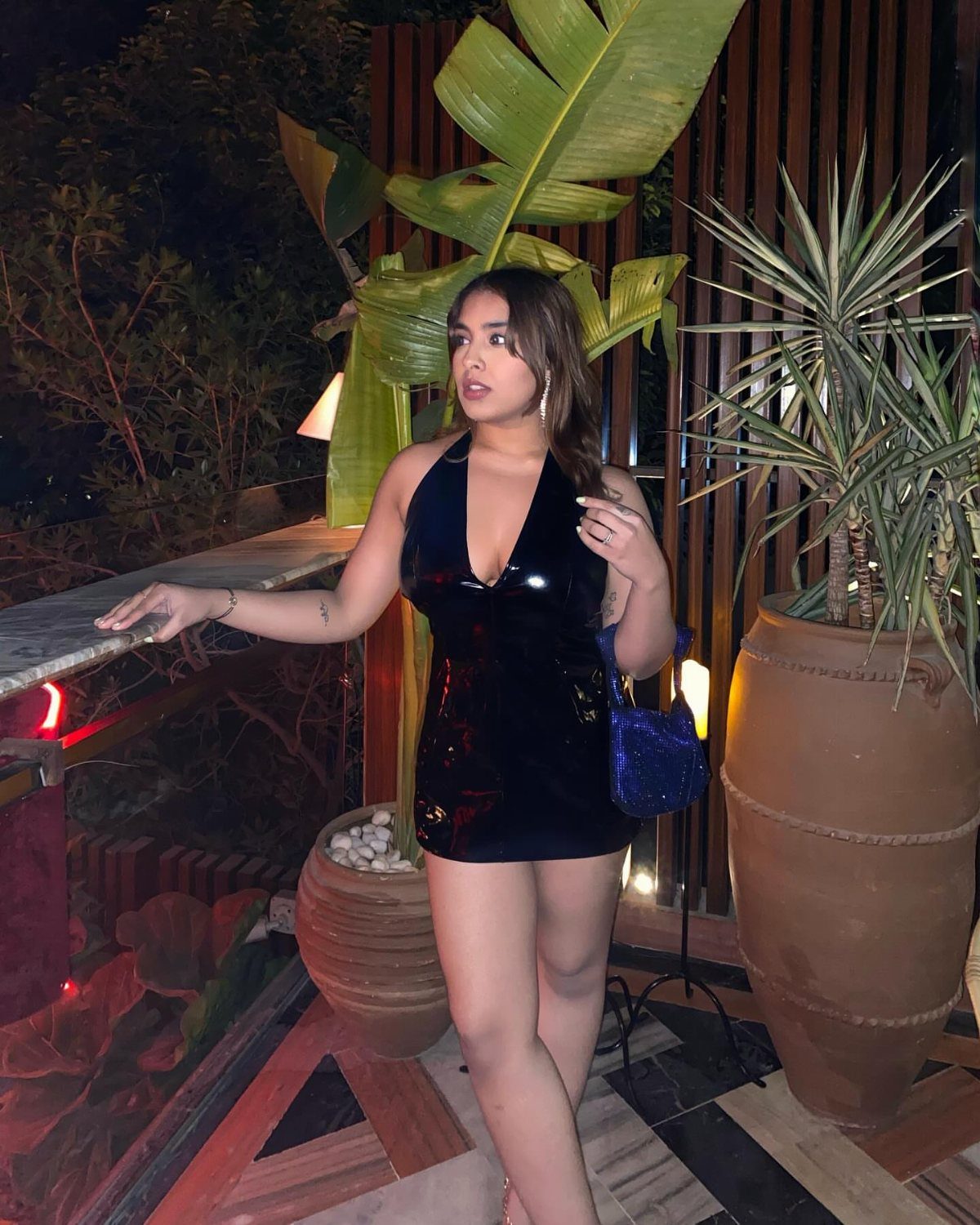 A woman posing in a short, shiny black satin dress on a balcony at night.