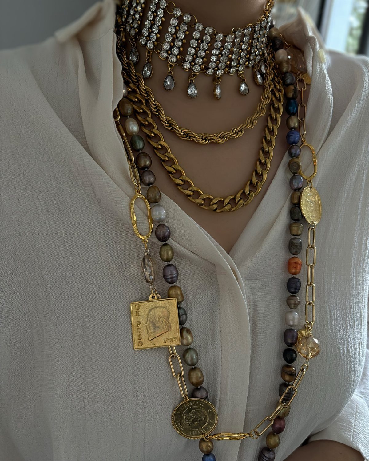 Multiple layered gold and rhinestone necklaces worn over a white linen shirt.