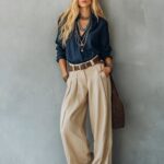 A blonde woman wearing a navy silk blouse and beige wide-leg trousers with layered necklaces.