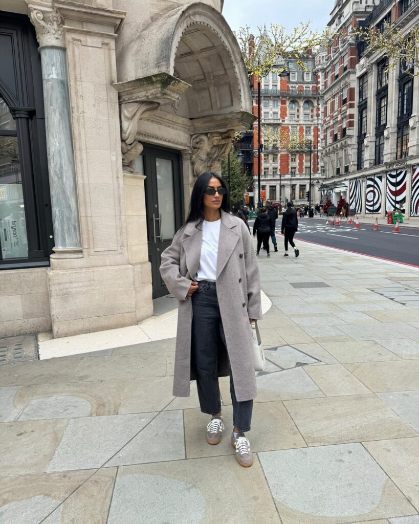 A woman wearing an oversized grey wool coat, white t-shirt, and black trousers standing on a city sidewalk.