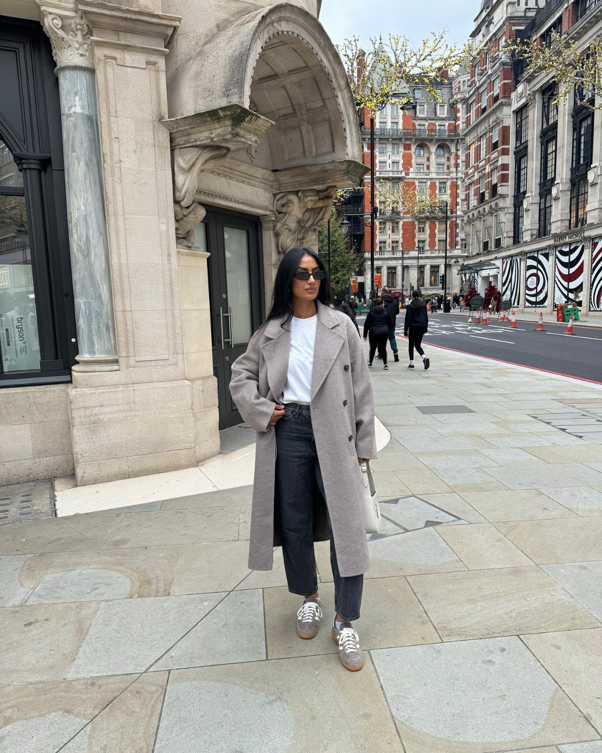 A woman wearing an oversized grey wool coat, white t-shirt, and black trousers standing on a city sidewalk.