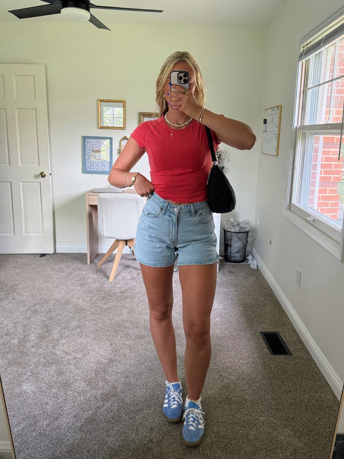 A woman posing in a red t-shirt and denim shorts for a mirror selfie.