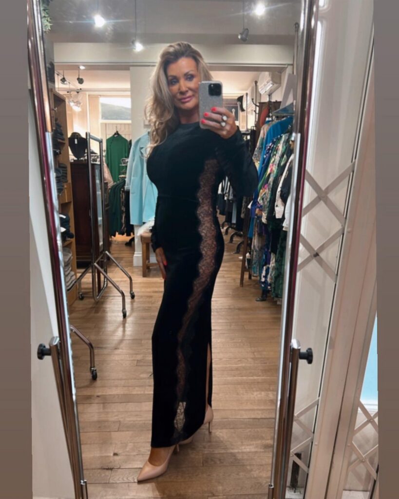 A woman in a long black lace dress posing for a mirror selfie in a boutique.