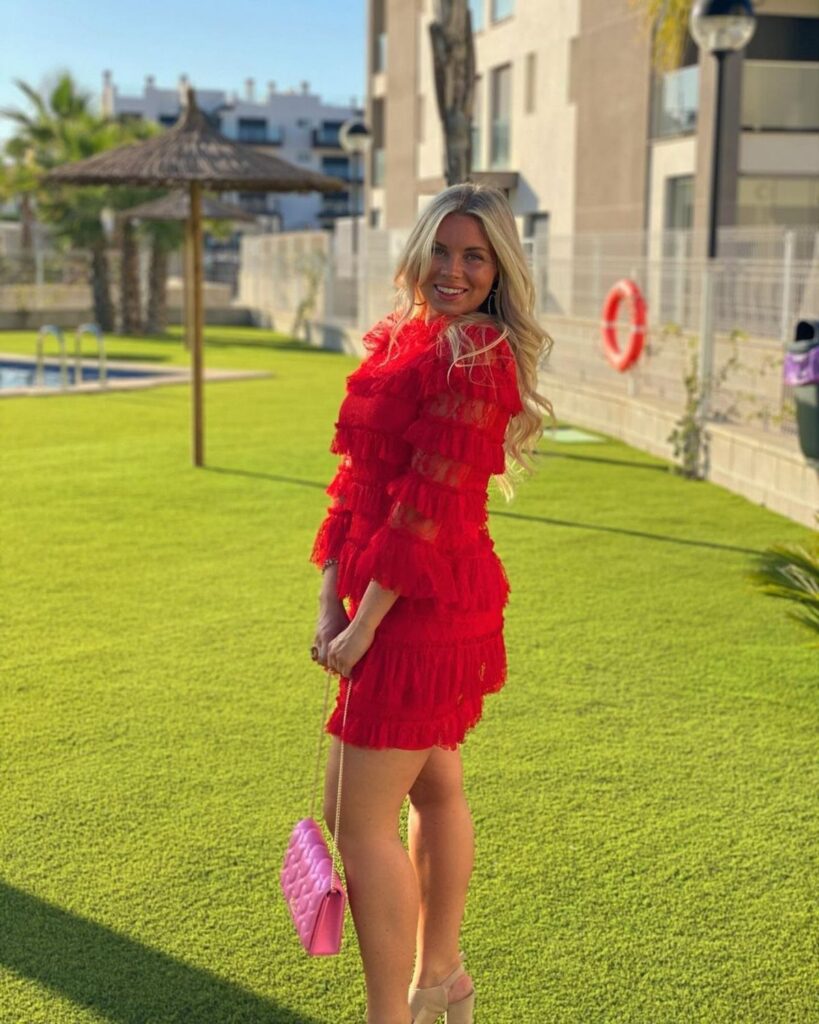 A smiling woman posing in a bright red tiered ruffled dress on a green lawn.