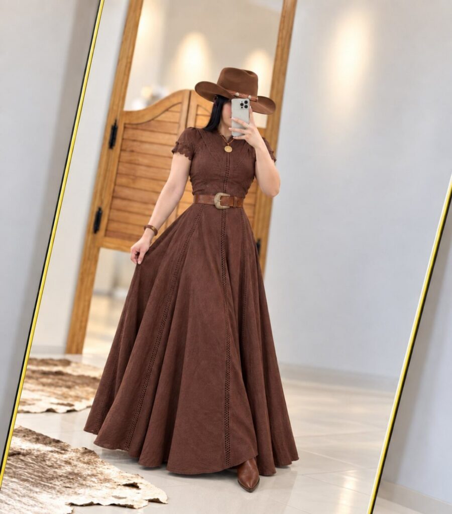 A woman in a long brown western dress and cowboy hat taking a mirror selfie.