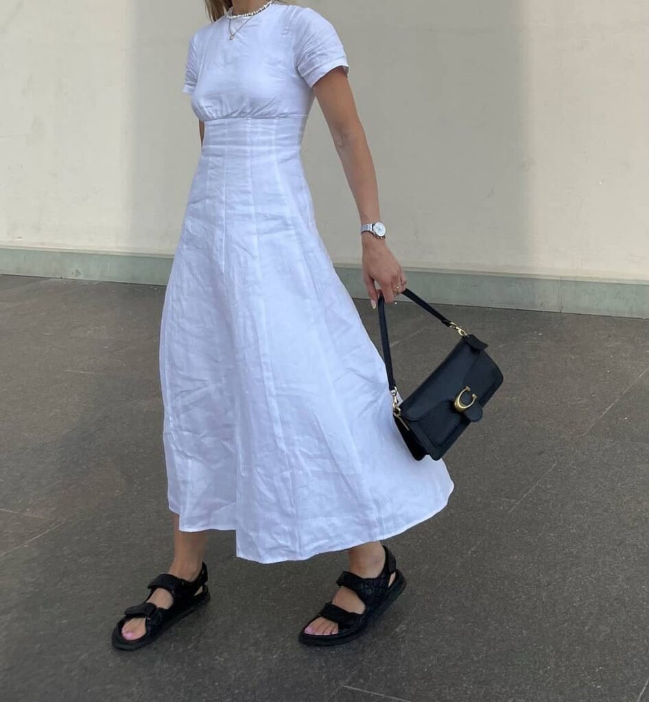 A person wearing a white short-sleeved midi dress, black sandals, and carrying a black handbag.