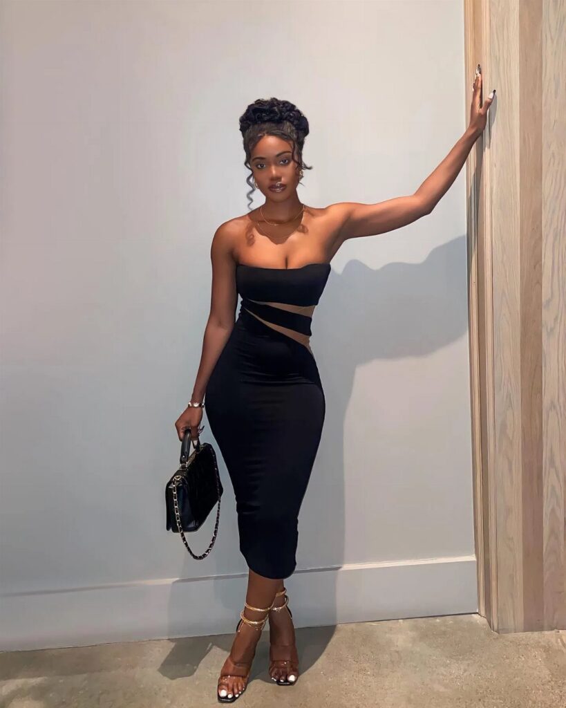 A woman in a black strapless body-con midi dress posing against a white wall.