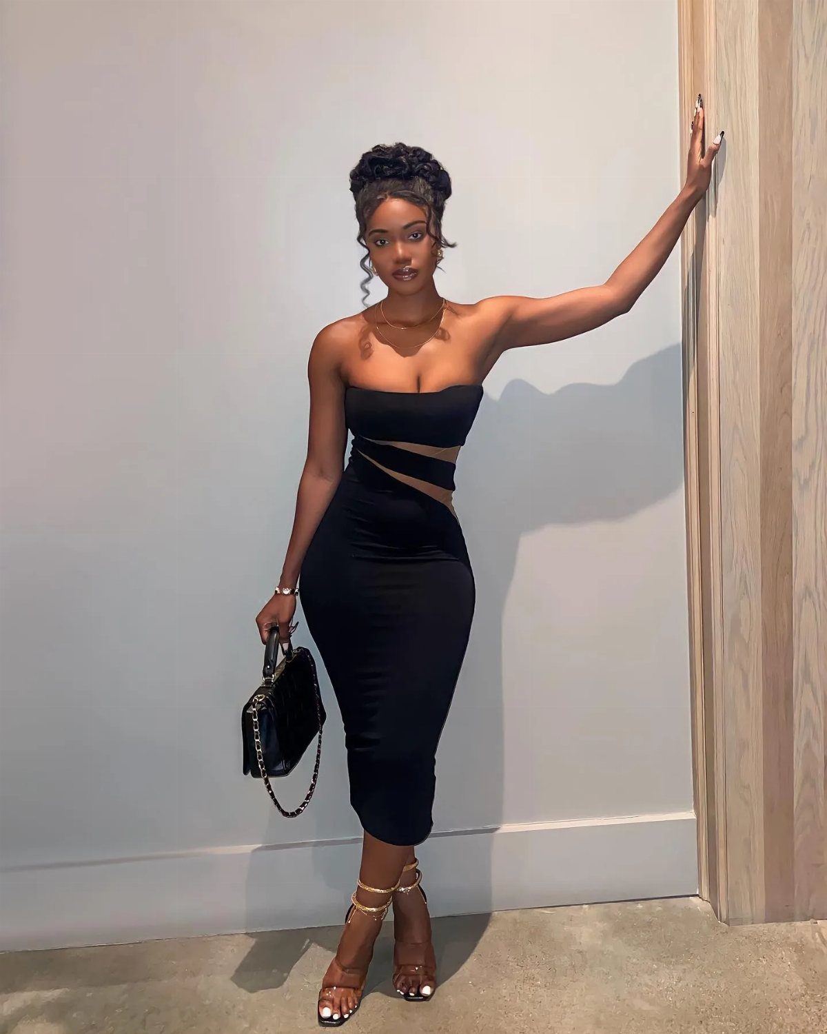 A woman in a black strapless body-con midi dress posing against a white wall.