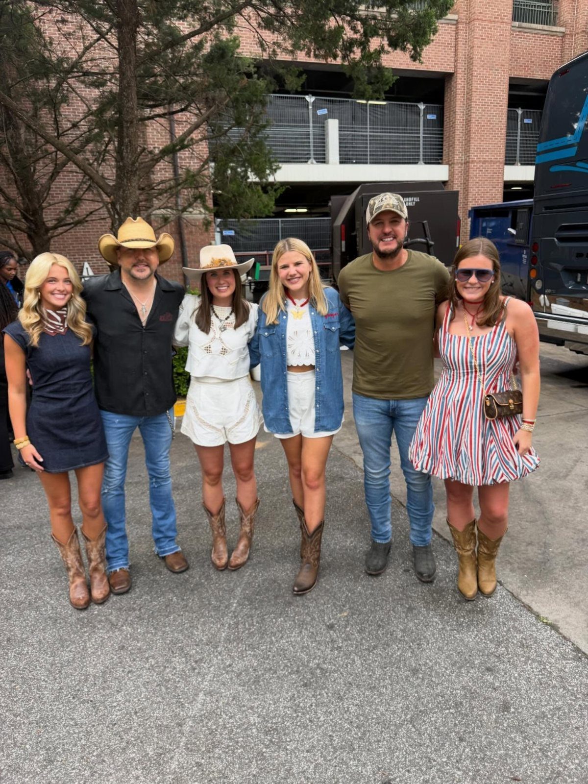 Six people posing together wearing various country-style outfits and cowboy boots outdoors.