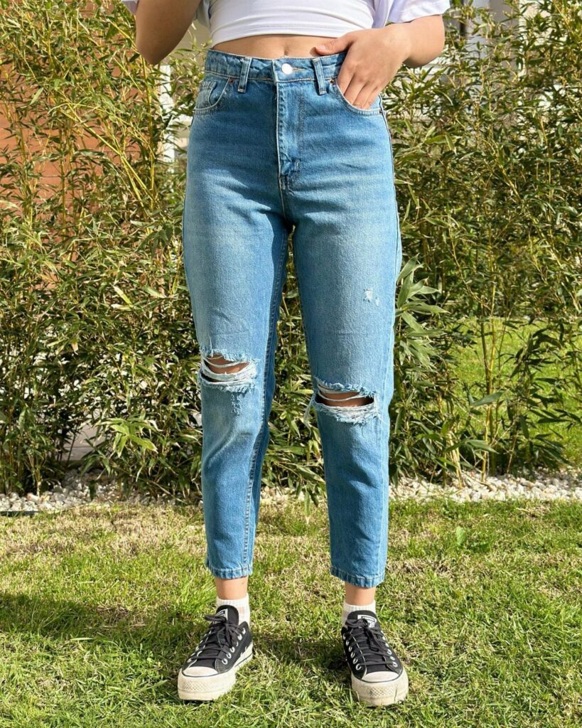 Person wearing high-waisted ripped blue jeans, a white crop top, and black sneakers outdoors.