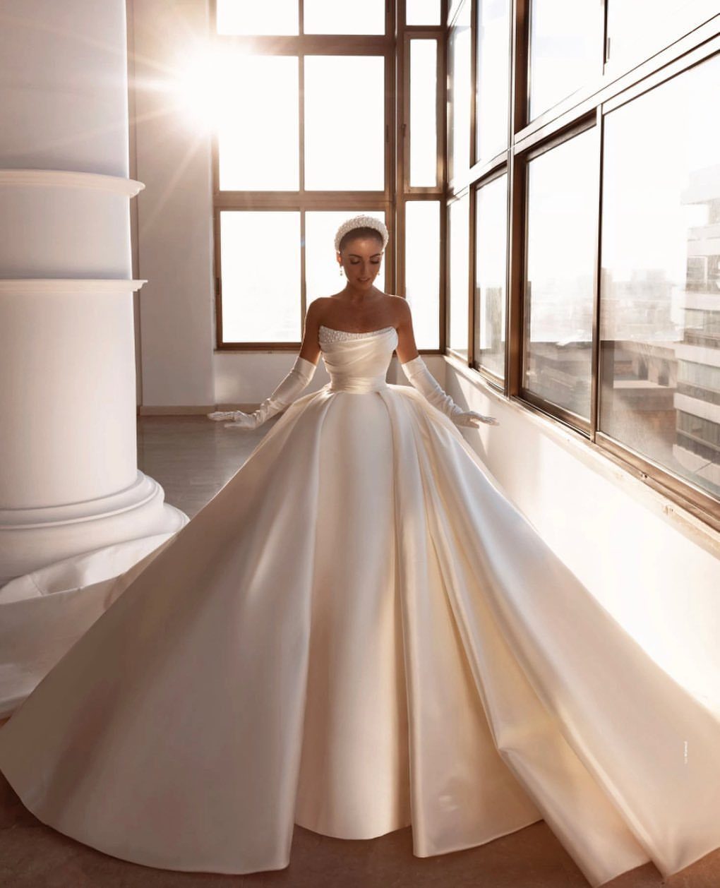 Woman wearing a voluminous white satin ballgown standing in a sunlit room by large windows.