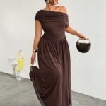 A woman wearing a long brown off-the-shoulder A-line dress holding a small handbag.