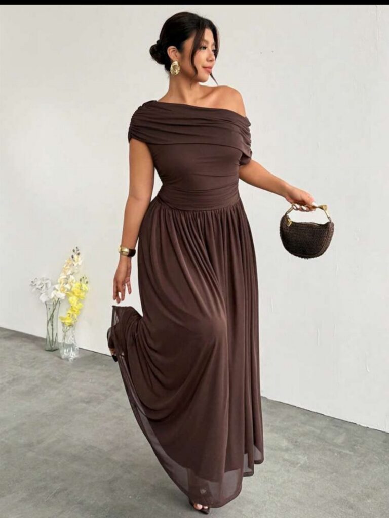 A woman wearing a long brown off-the-shoulder A-line dress holding a small handbag.