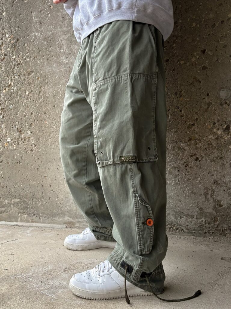 Olive green baggy cargo pants with large pockets paired with white sneakers against a concrete wall.