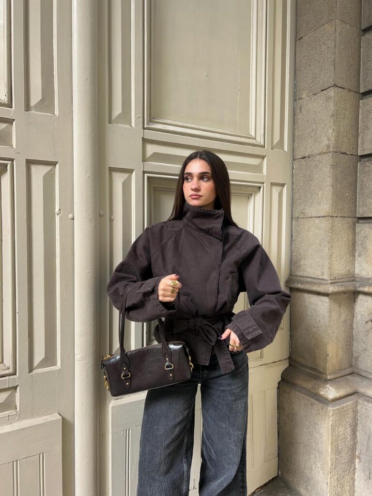 A woman wearing a dark brown funnel-neck jacket and wide-leg jeans standing by a door.