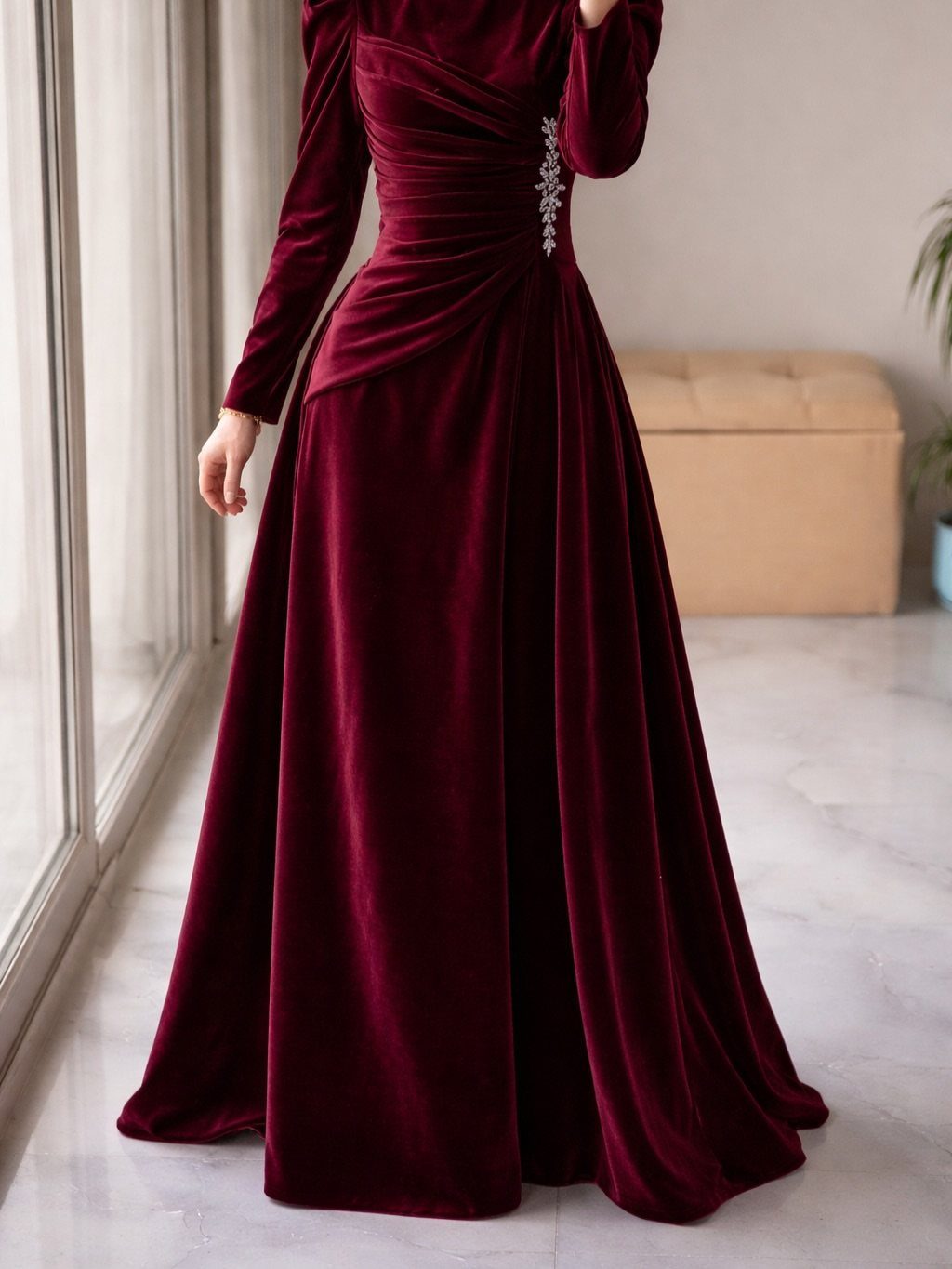 A person wearing a long-sleeved, floor-length burgundy velvet gown with ruched waist detailing.