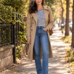 A woman wearing a striped Breton top, blue straight-leg jeans, and a tan trench coat outdoors.