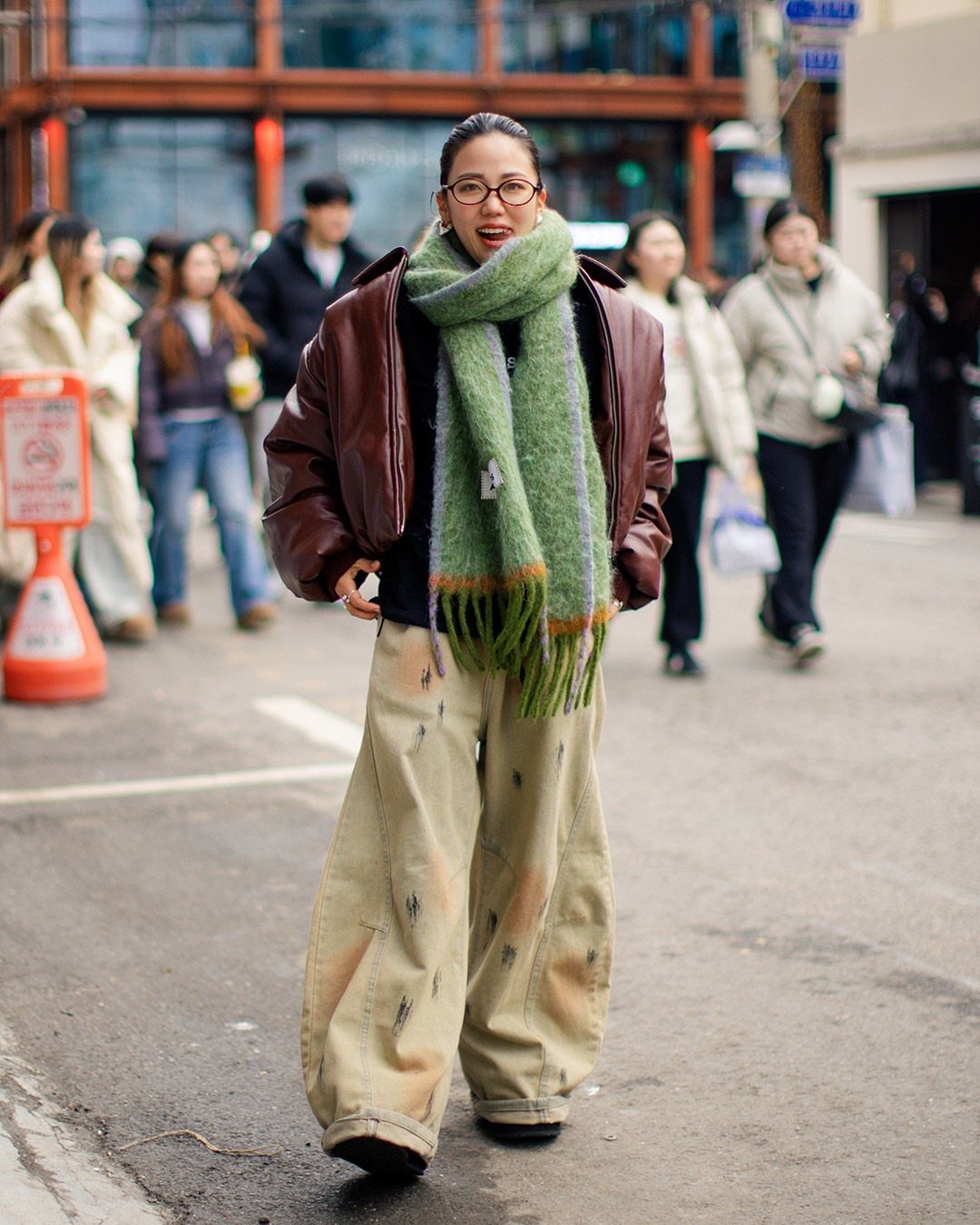 A woman in a leather jacket and green scarf walking through a busy city street.