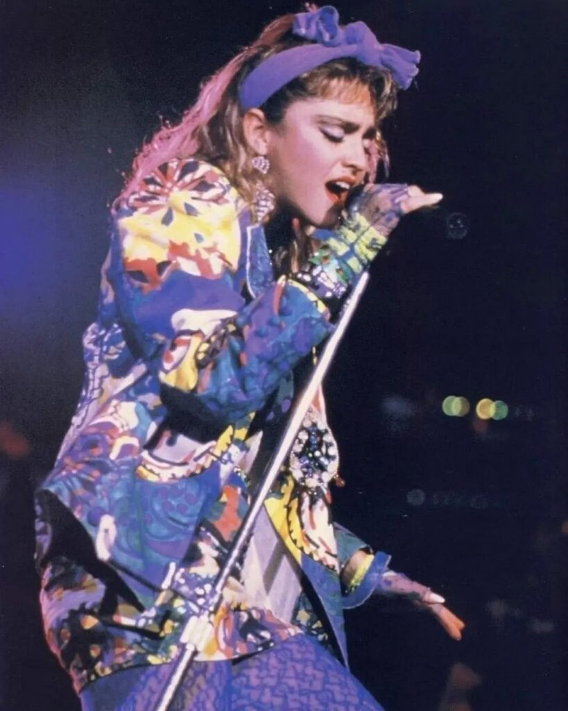 Madonna singing on stage wearing a colorful patterned jacket and a purple headband.