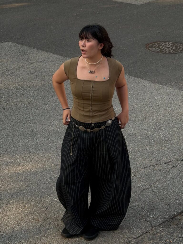 A woman in an olive corset and black pinstriped wide-leg trousers standing on an asphalt street.