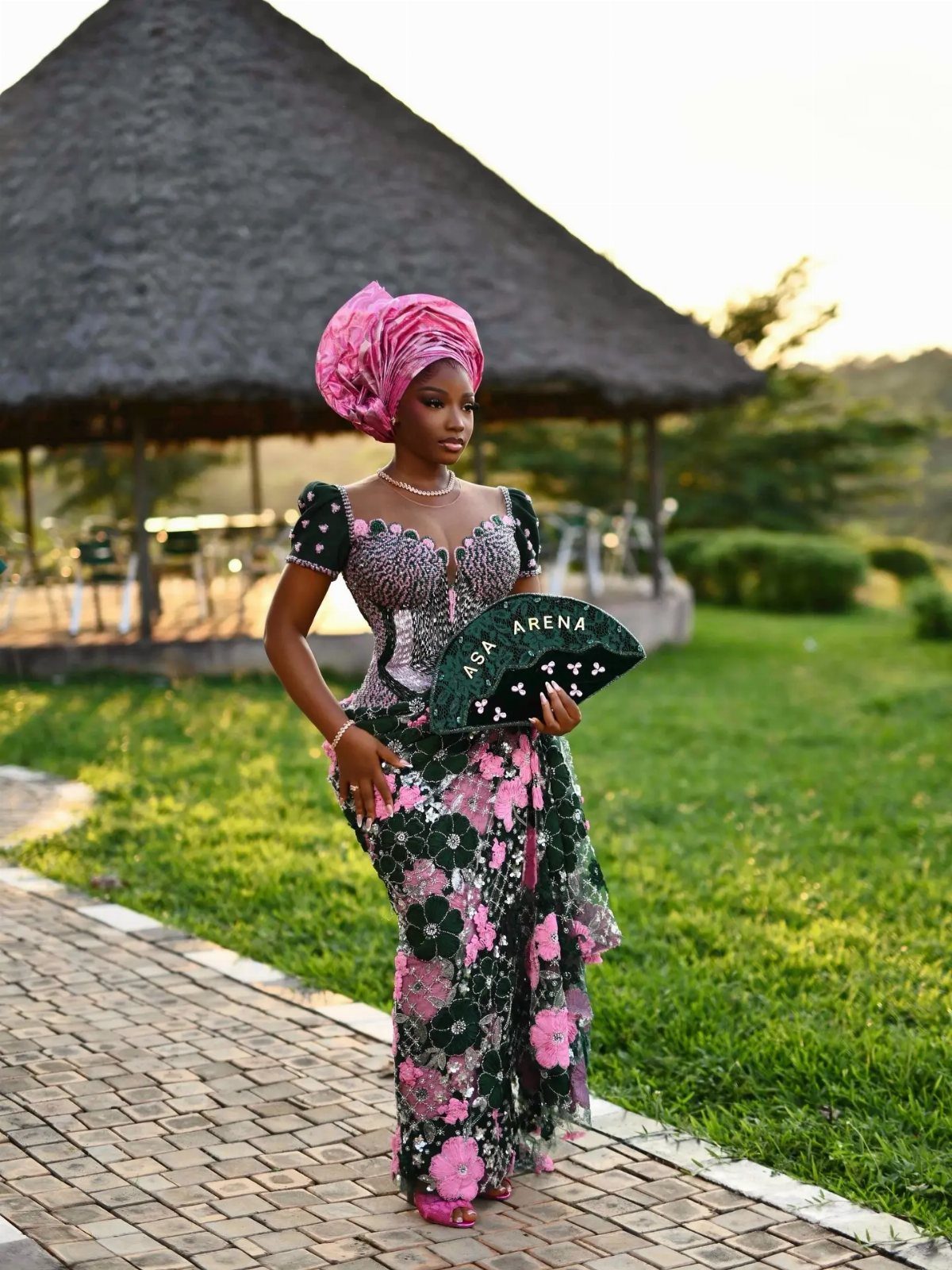 A woman in traditional Nigerian lace attire and a pink gele headwrap posing outdoors.