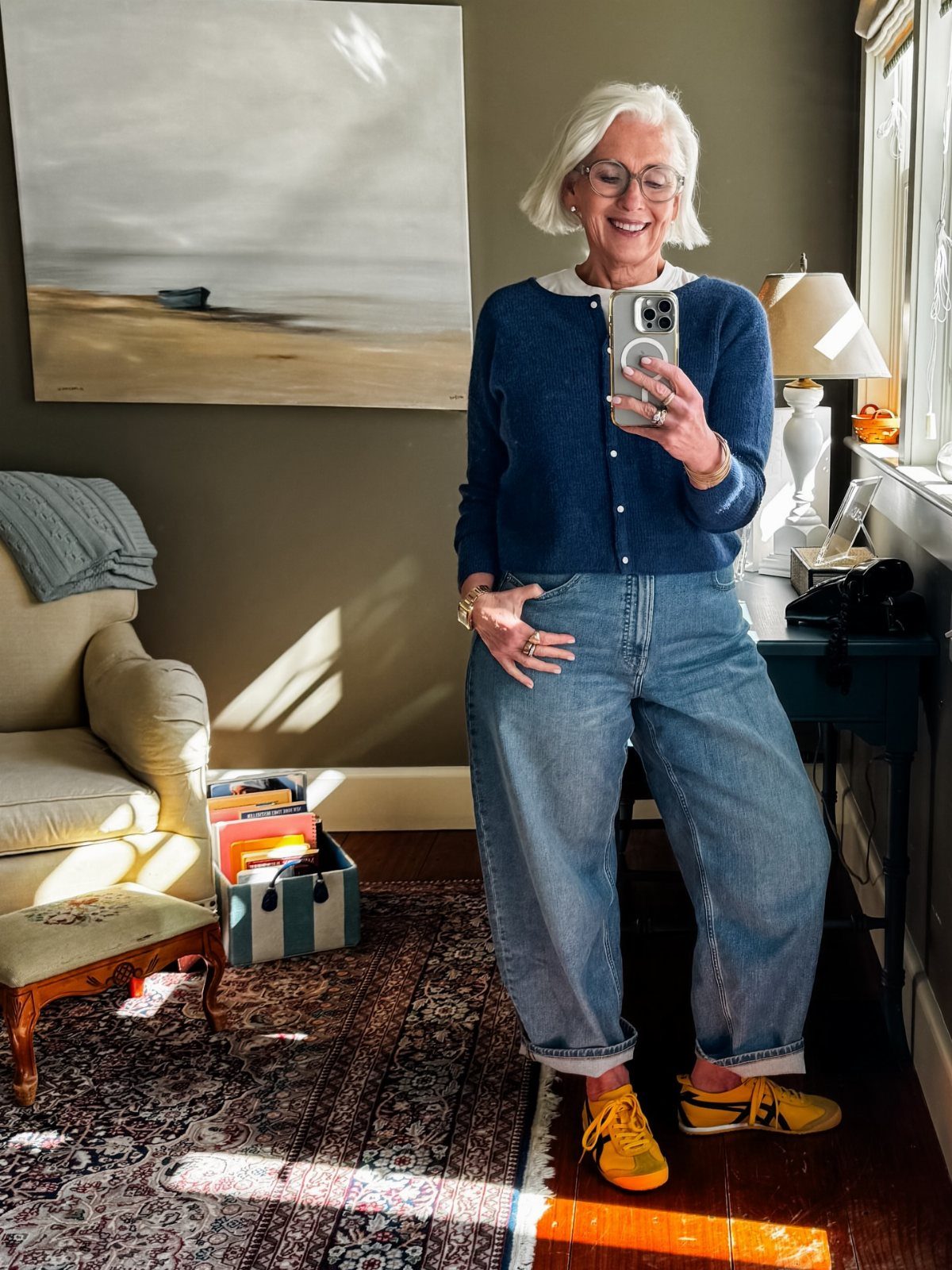 A smiling older woman with white hair posing in a blue cardigan and baggy jeans.