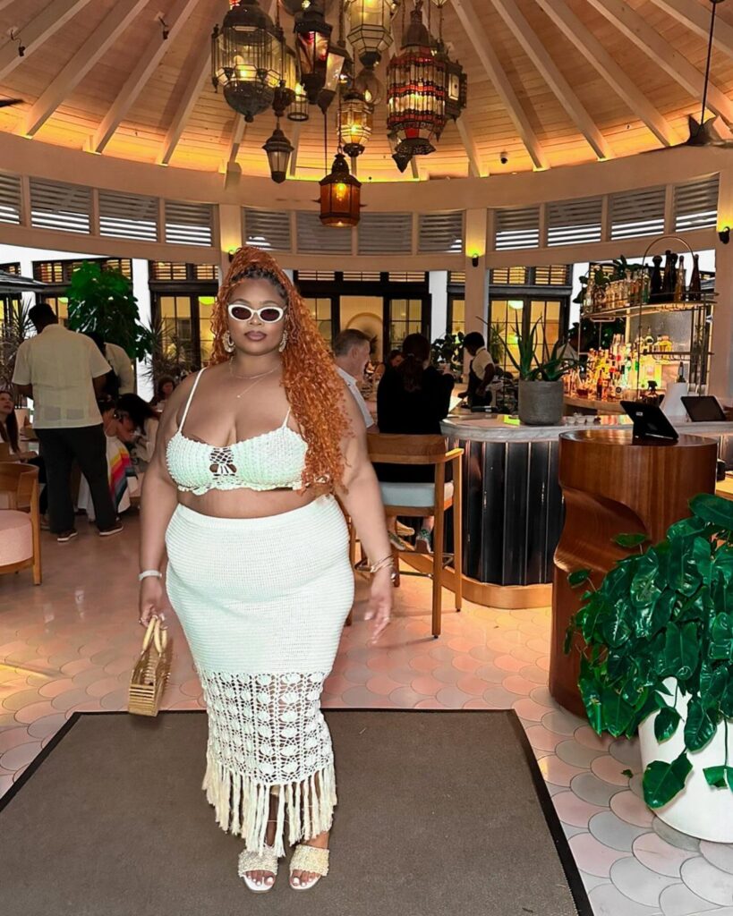 A plus size woman posing in a white crochet two-piece outfit at a tropical lounge.