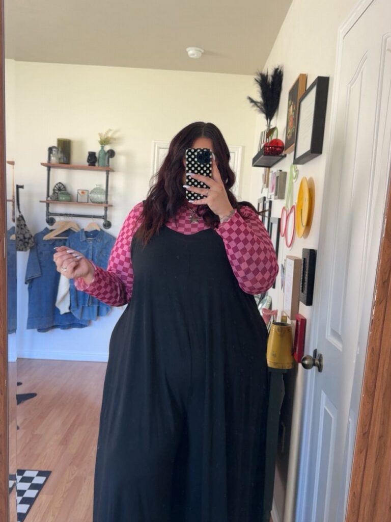 A person wearing pink patterned sleeves and black wide-leg overalls taking a mirror selfie.