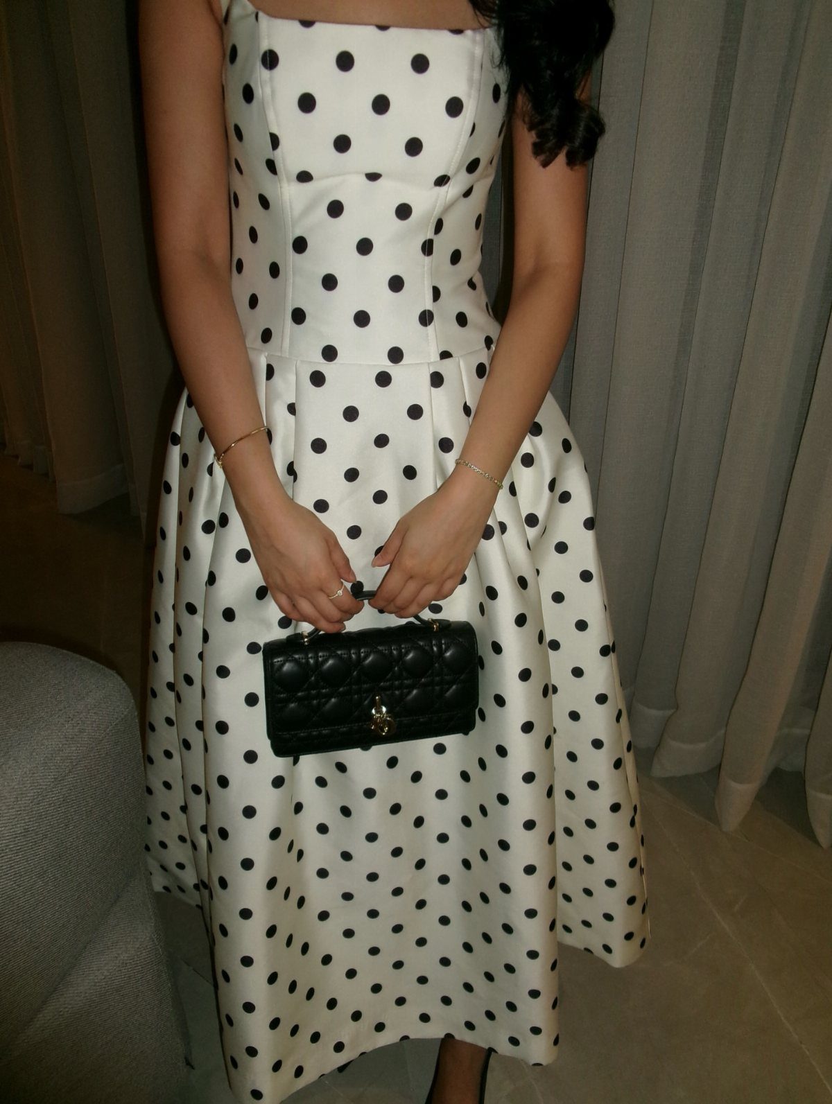 A person wearing a white midi dress with large black polka dots and holding a black handbag.