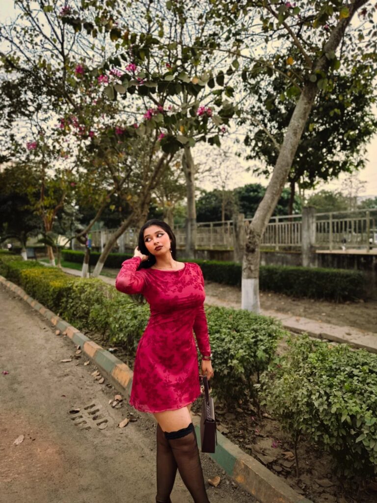 A woman in a red patterned dress and black stockings posing outdoors near green hedges.