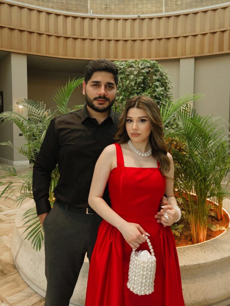 A woman wearing a vibrant red dress and pearl accessories standing next to a man.