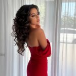 A woman with long dark curly hair wearing a red off-the-shoulder dress.
