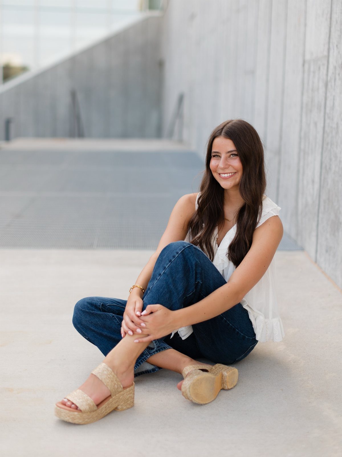 A smiling young woman sitting on concrete wearing a white blouse and blue jeans.