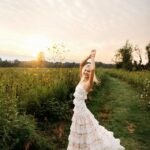 Blonde woman posing in a white floral tiered maxi dress in a grassy field at sunset.