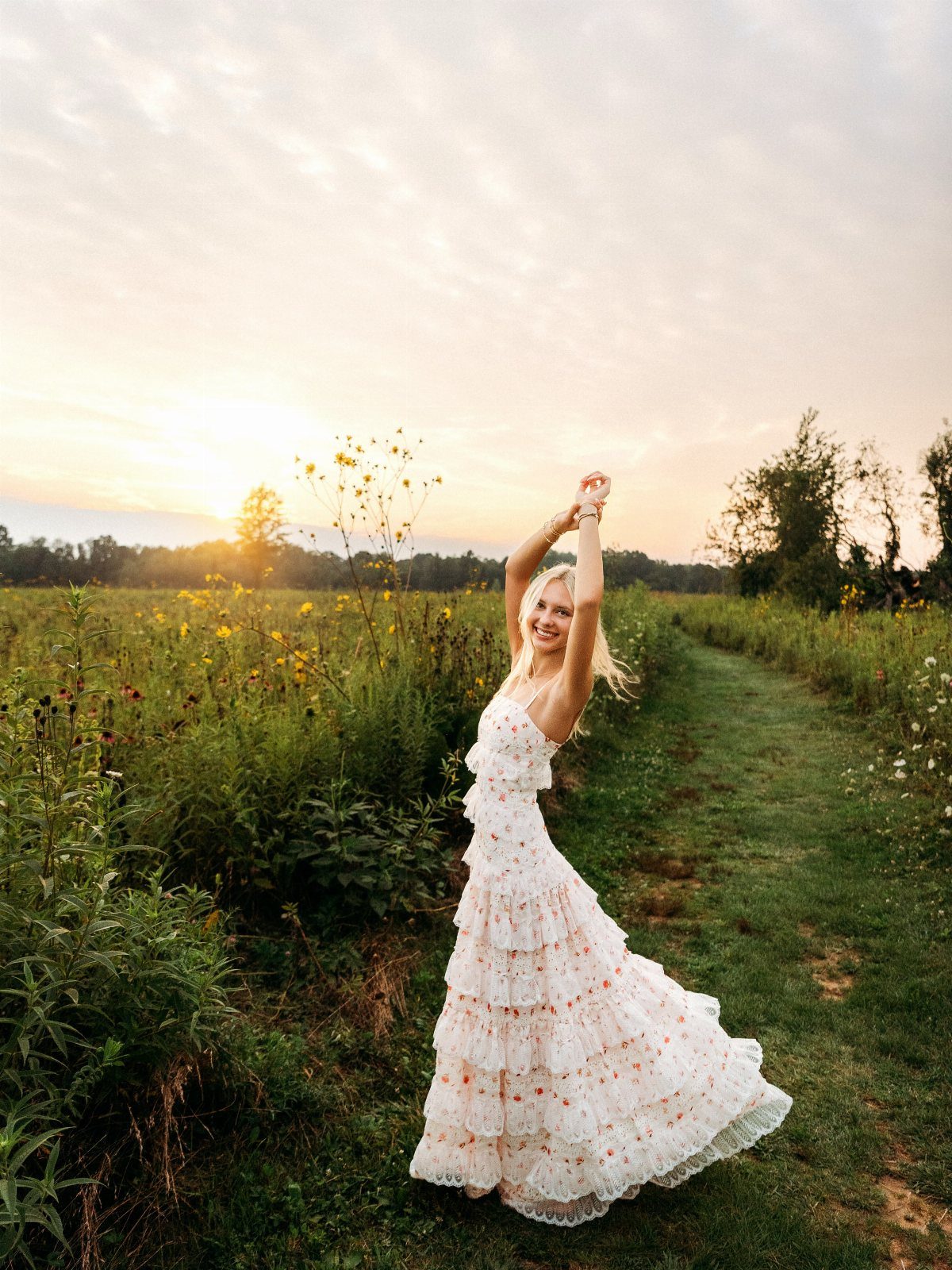 Blonde woman posing in a white floral tiered maxi dress in a grassy field at sunset.