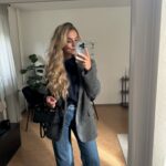 A blonde woman posing in a mirror wearing a grey blazer, navy top, and blue jeans.