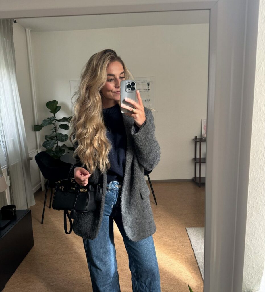 A blonde woman posing in a mirror wearing a grey blazer, navy top, and blue jeans.