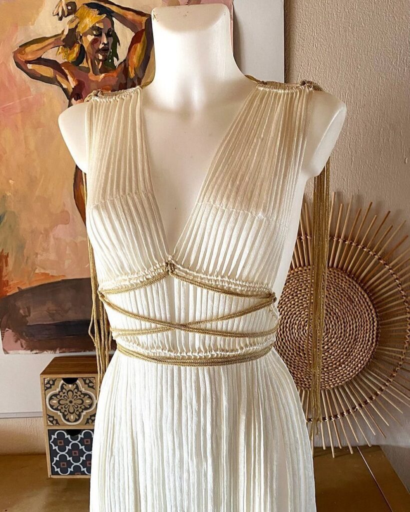 White pleated dress with deep V-neckline and gold cord accents on a mannequin.