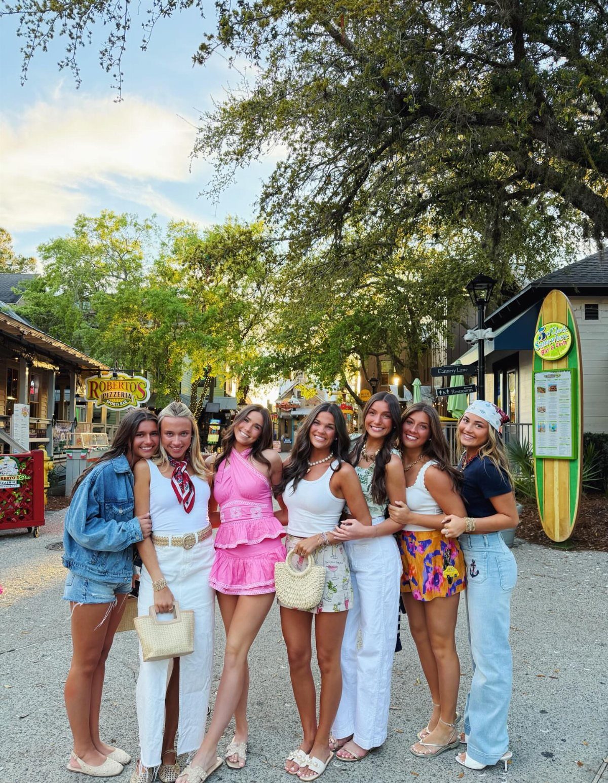 Seven young women posing together in various colorful summer vacation outfits outdoors.
