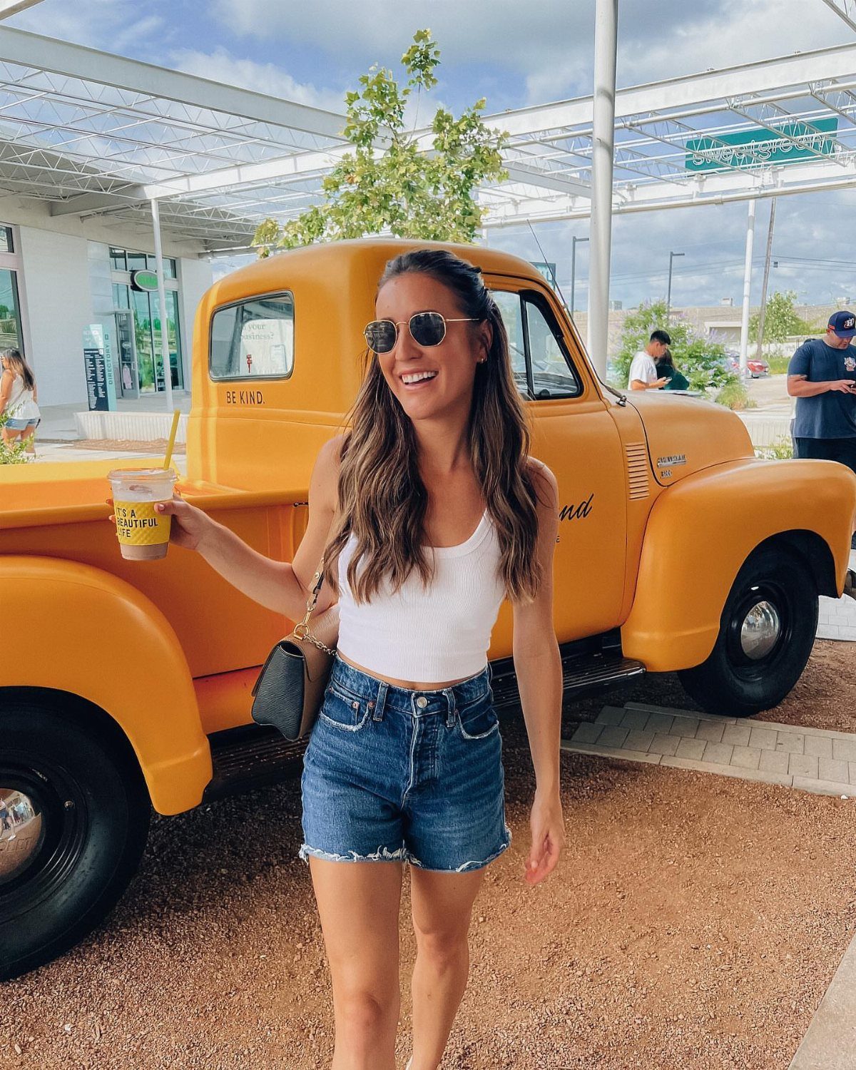A smiling woman in a white tank top and denim shorts standing by a yellow truck.