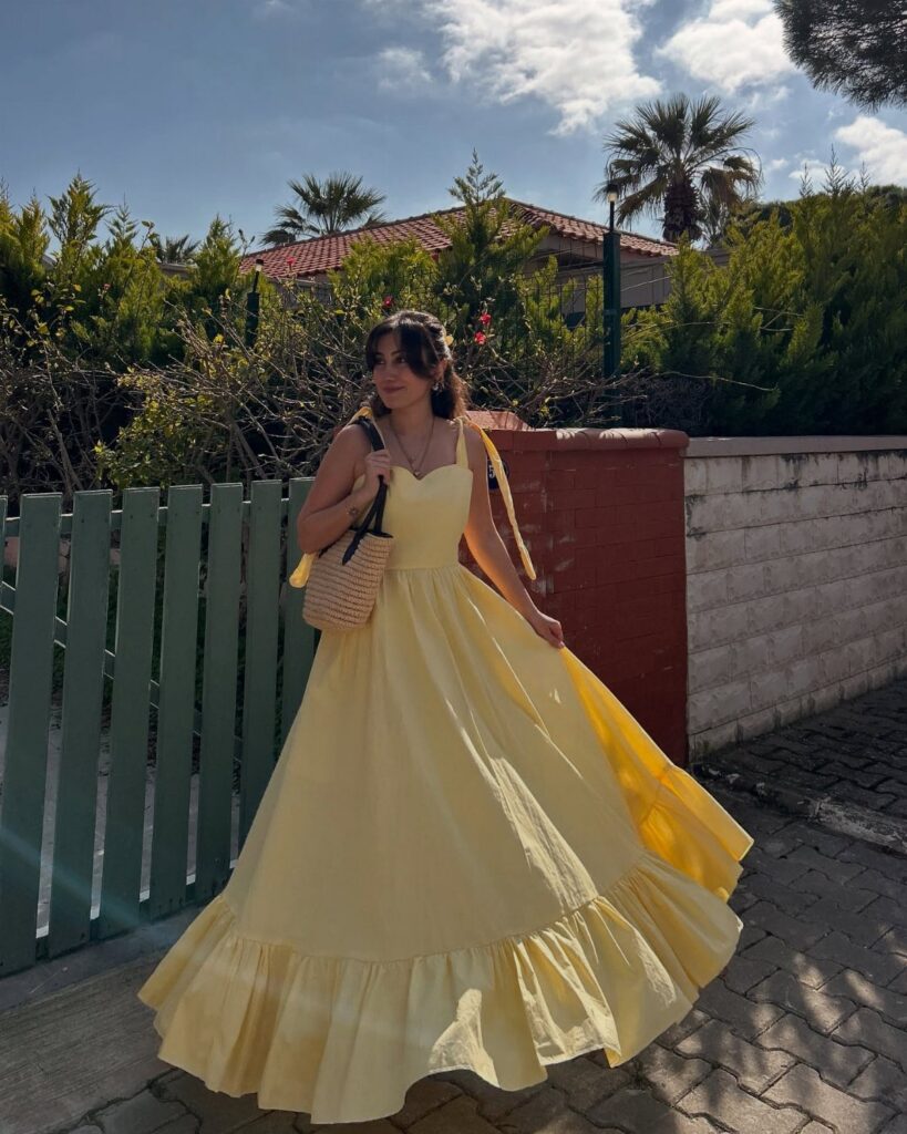 Woman in a long, pale yellow sleeveless maxi dress standing by a green fence.